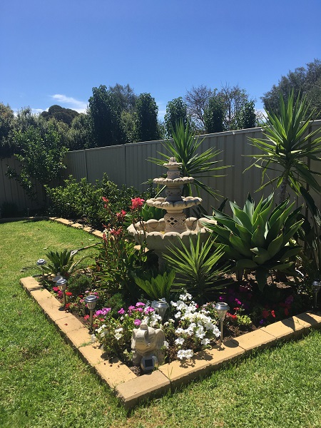 A white five tier water fountain in a garden bed with flowersand plants