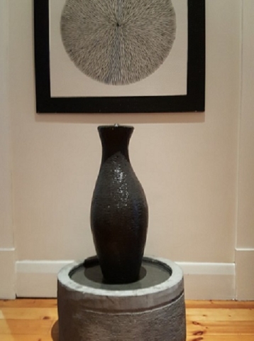A small organic shaped urn water feature is positioned indoors with an abstract painting on the wall behind it